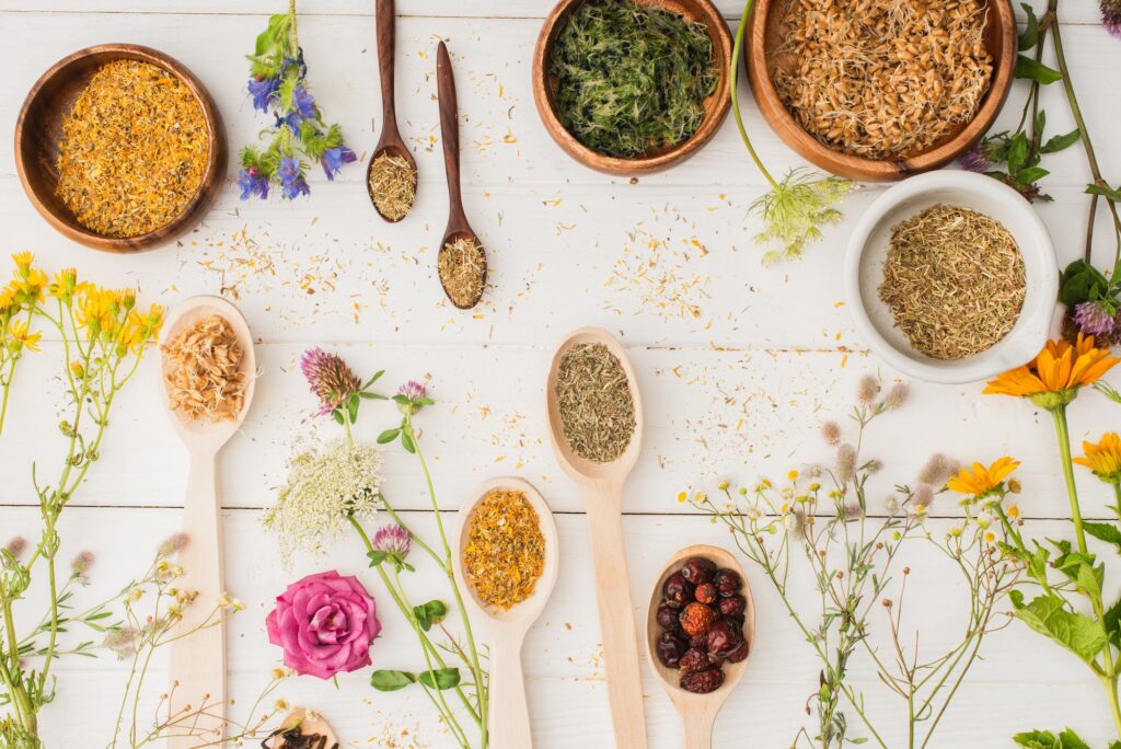 top view of herbs in spoons and bowls near flowers on white wooden background naturopathy concept