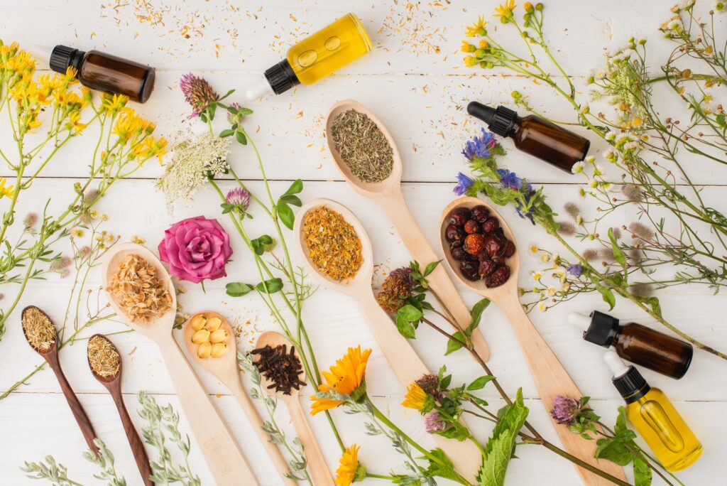 top view of herbs in spoons near flowers and bottles on white wooden background naturopathy concept